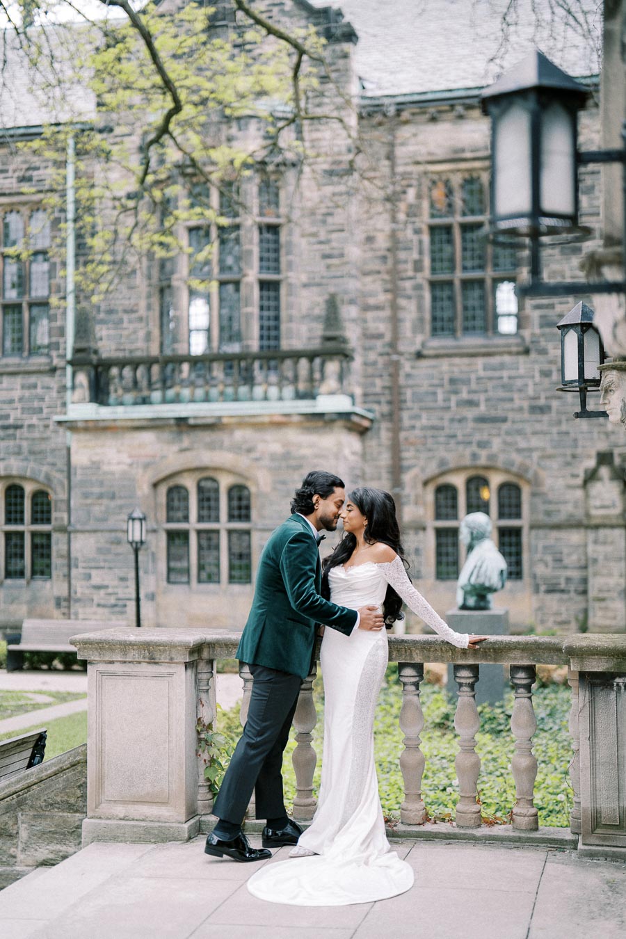A romantic couple embraces in front of a historic stone building, with the woman in a long, white dress and the man in a dark suit, surrounded by elegant architecture and lush greenery.