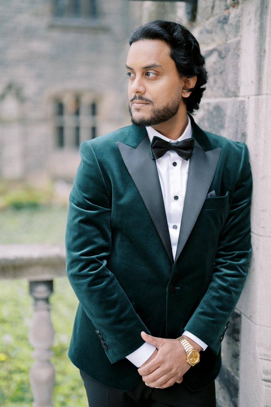 A person in a green velvet tuxedo and bow tie standing outdoors next to a stone wall.