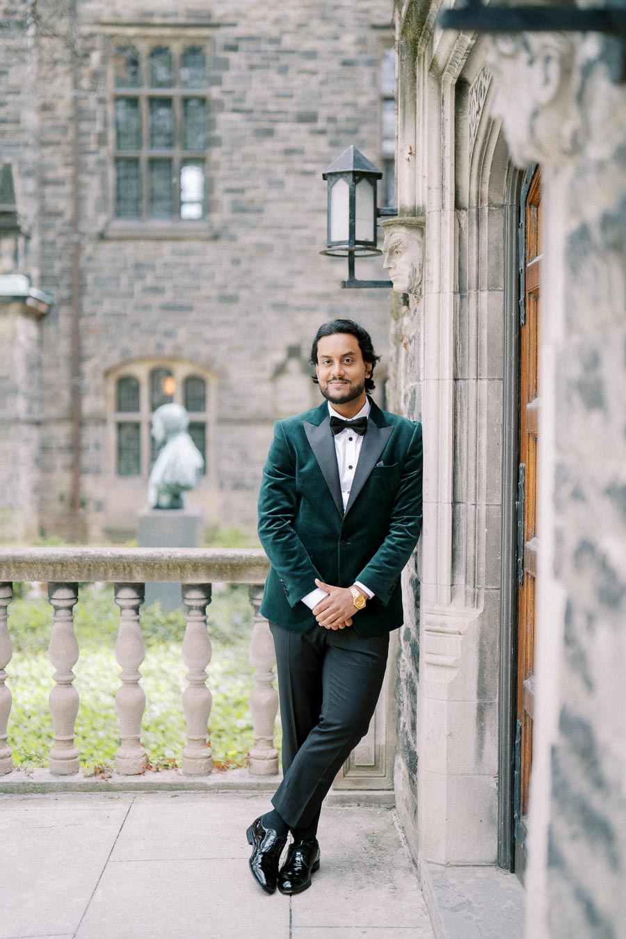 Man in a dark green suit with a black bow tie leaning casually against a stone building, with a historic architectural backdrop.