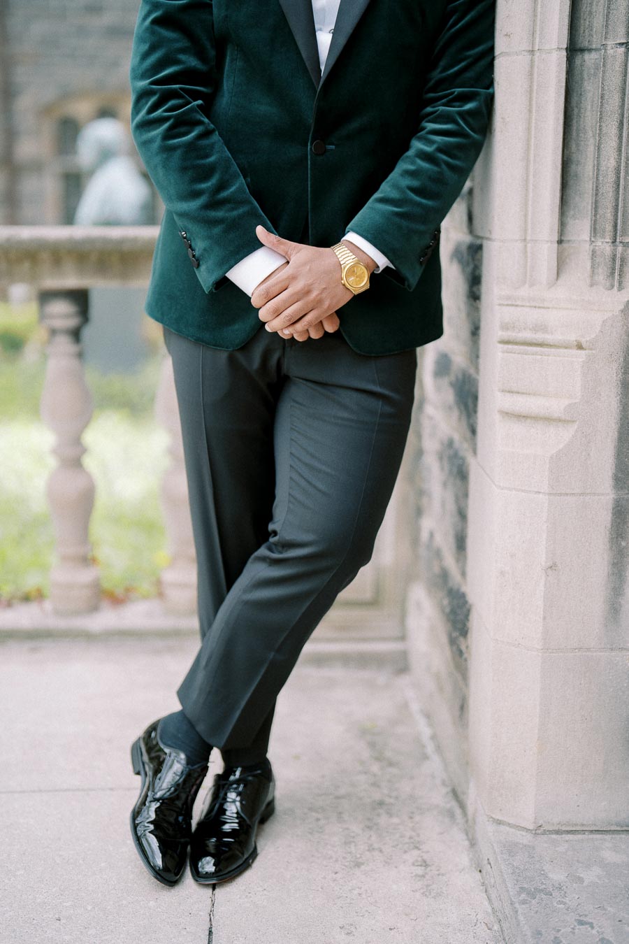 A well-dressed man in a dark green velvet blazer and black pants stands casually against a stone wall. He is wearing shiny black shoes and a gold watch, with hands clasped together. The setting appears to be outdoors, with a blurred garden and stone railing in the background.