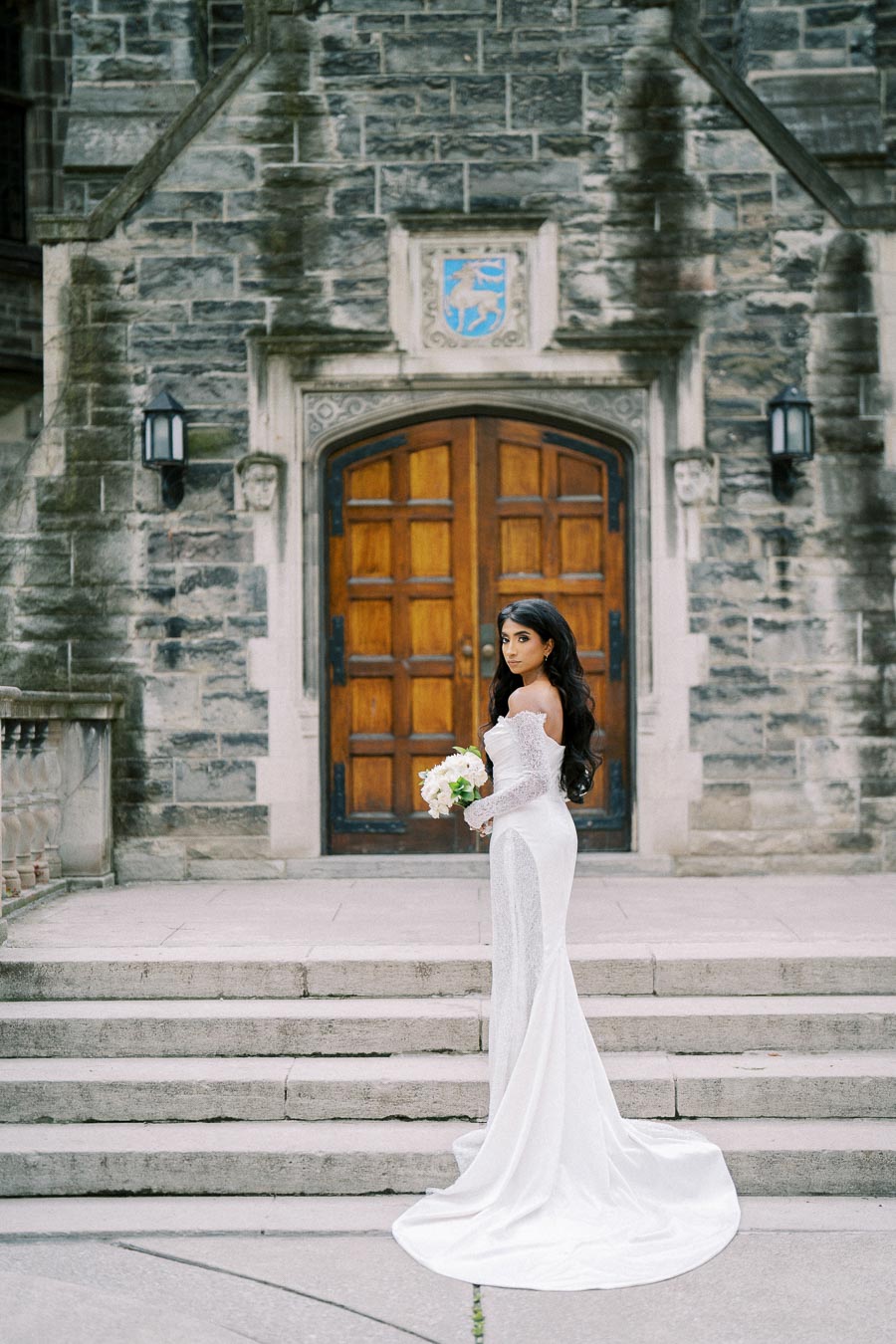 A bride in an elegant white gown poses on stone steps in front of a historic building with a wooden door and ornate architecture, holding a bouquet of white flowers.