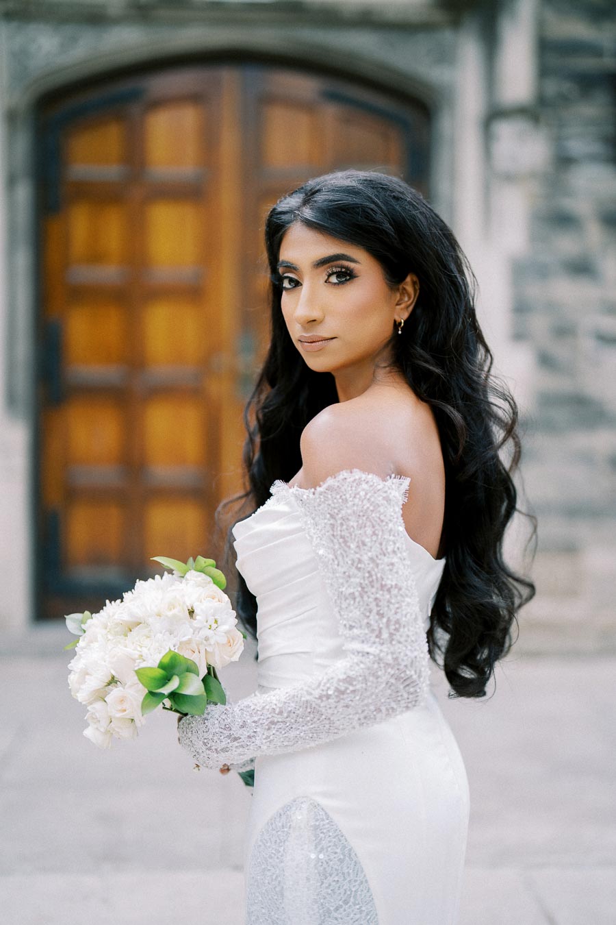 A bride in an elegant white gown holding a bouquet of white flowers stands in front of an ornate wooden door, with a captivating gaze. The setting is a stone-walled building, adding a timeless and romantic atmosphere.