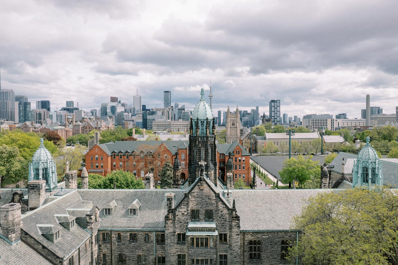 Aerial view of a historic university campus with old stone buildings and a scenic urban skyline in the background under a cloudy sky.