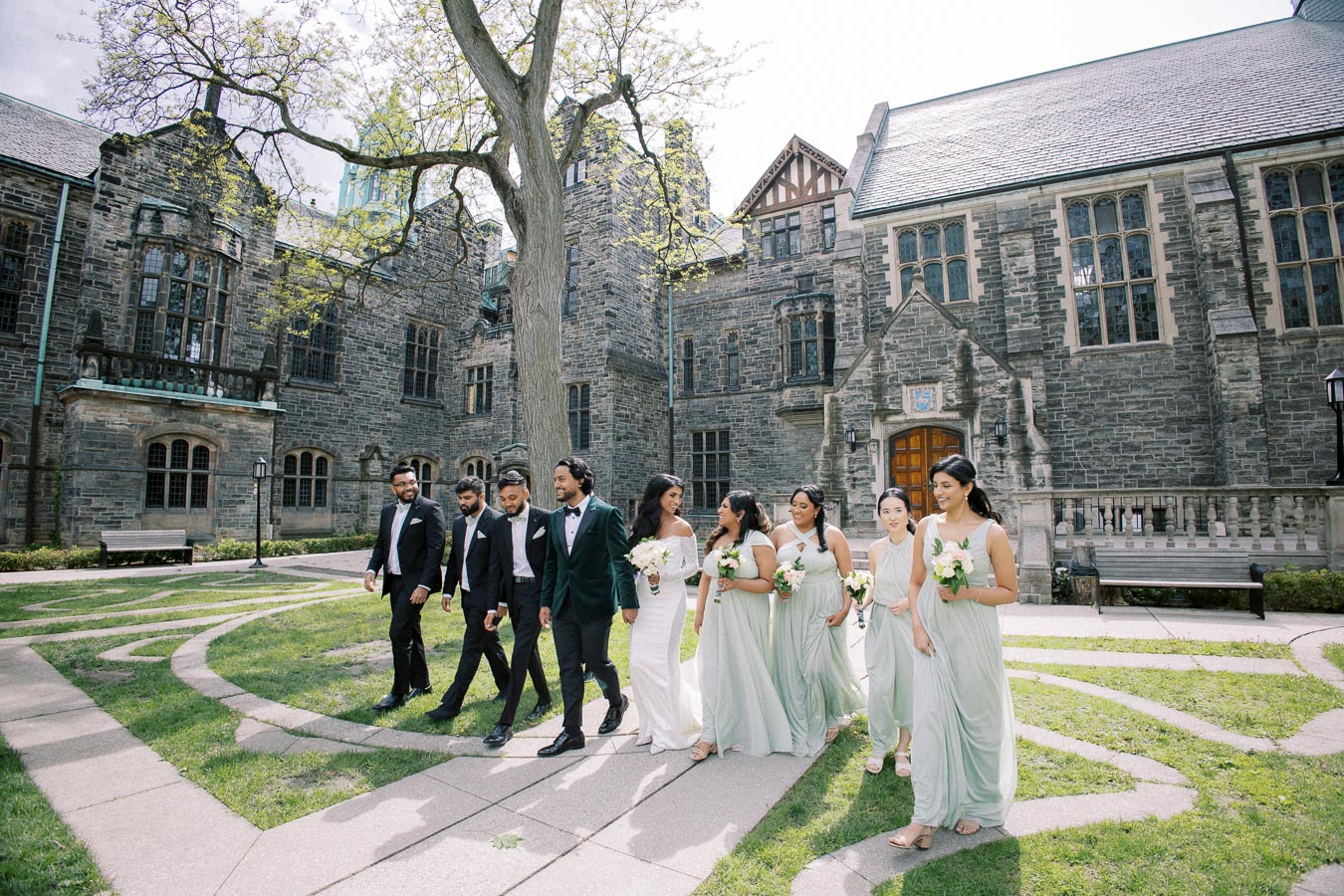 A wedding party walking in front of a historic stone building, with bridesmaids in light green dresses and groomsmen in dark suits, enjoying a sunny day surrounded by greenery.