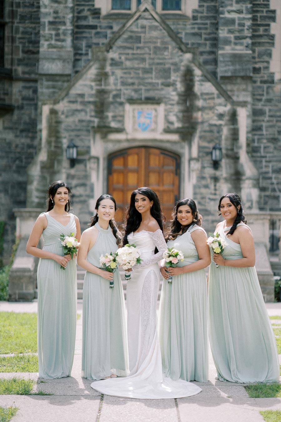 Bride in an elegant white dress poses with bridesmaids in mint green gowns holding bouquets in front of a stone building, capturing a beautiful wedding day moment.