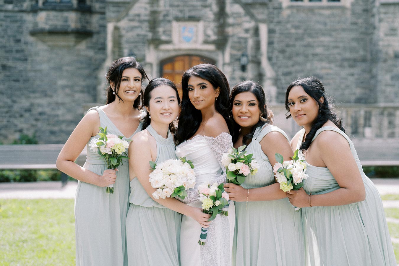 A bride in a white gown with bridesmaids in matching light green dresses, holding bouquets of white and pink flowers, posing happily in front of a historic stone building on a sunny day.