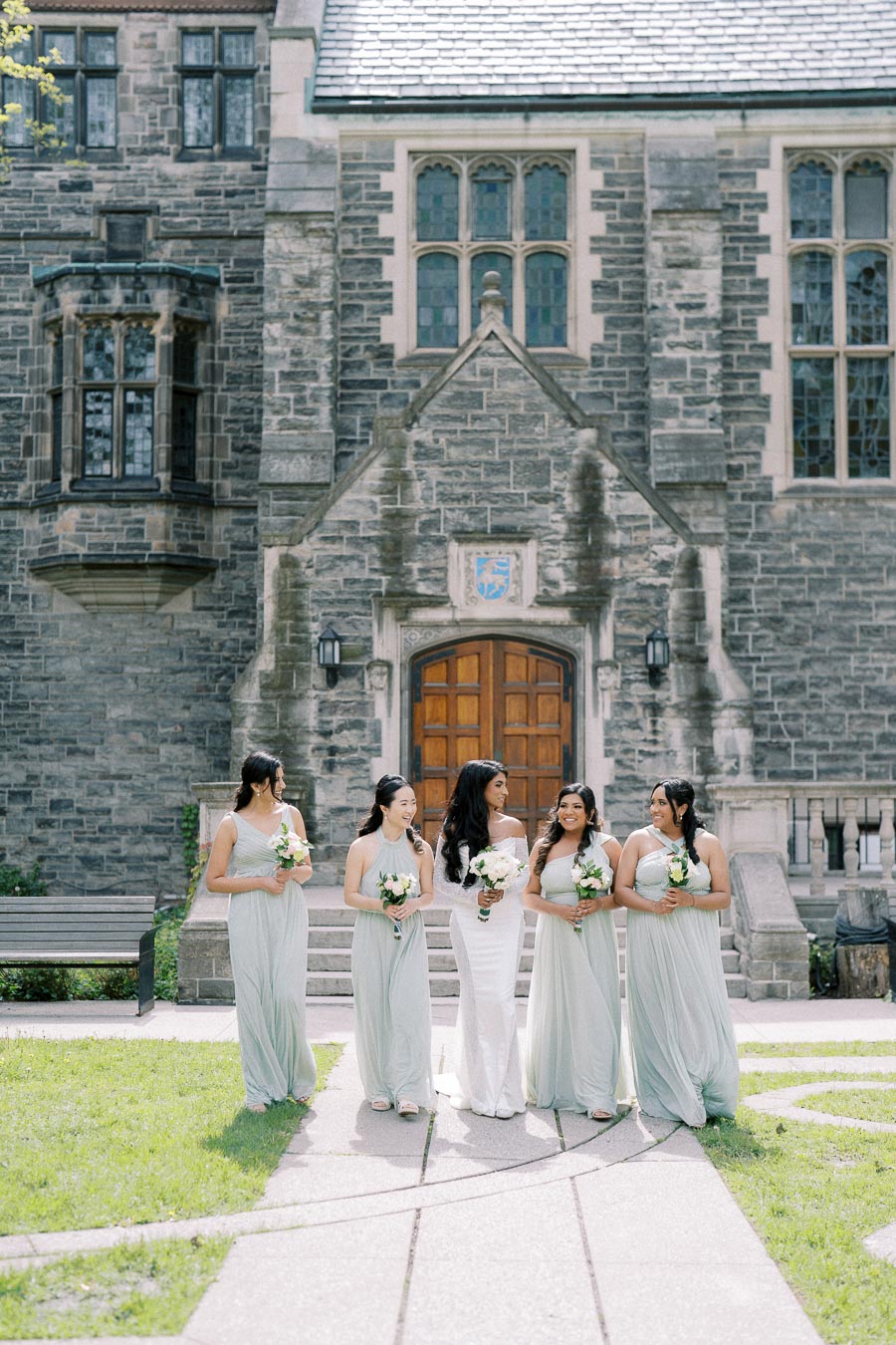 A bride in a white gown standing with four bridesmaids in light green dresses, each holding bouquets, in front of a historic stone building with arched windows and a wooden door.