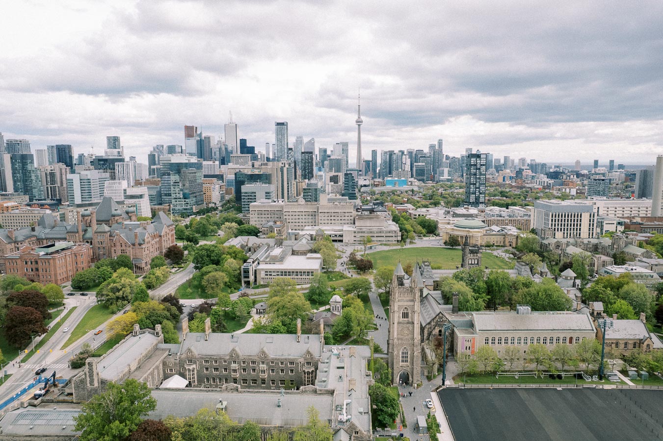 Aerial view of a university campus with historic buildings surrounded by lush greenery in the foreground, set against the backdrop of a modern city skyline under a cloudy sky.