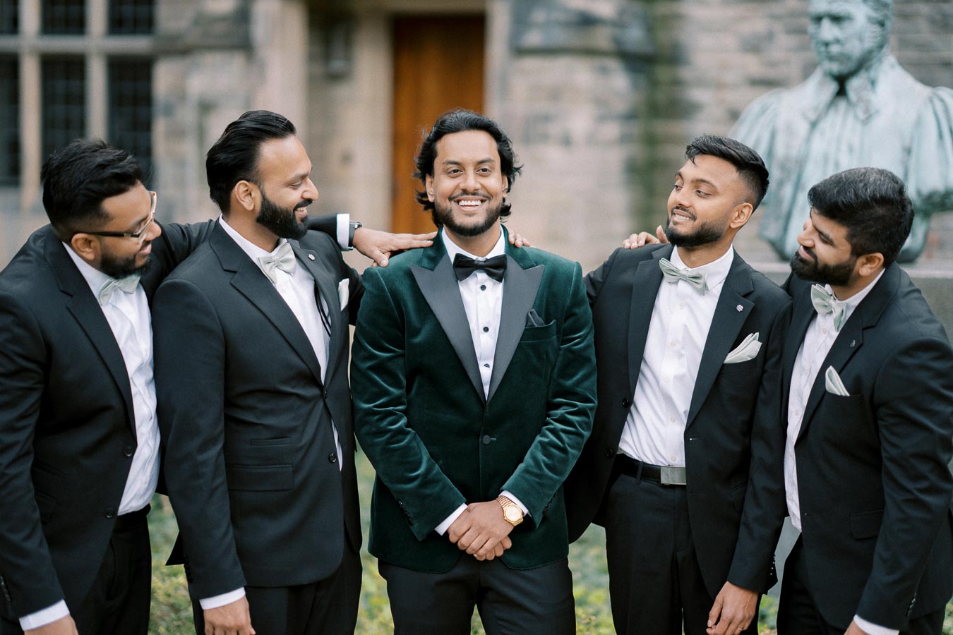 A group of five men dressed in formal attire, including a man in a green velvet tuxedo, smiling and standing together in an outdoor setting with a stone building in the background.
