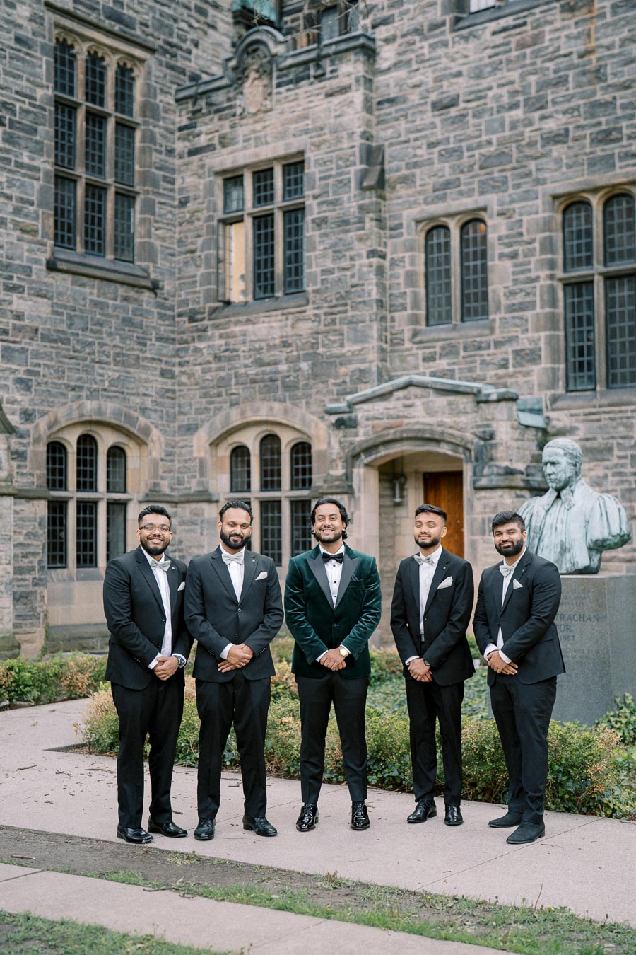 Five men in formal suits stand smiling in front of a historic stone building, showcasing a classic architectural background.