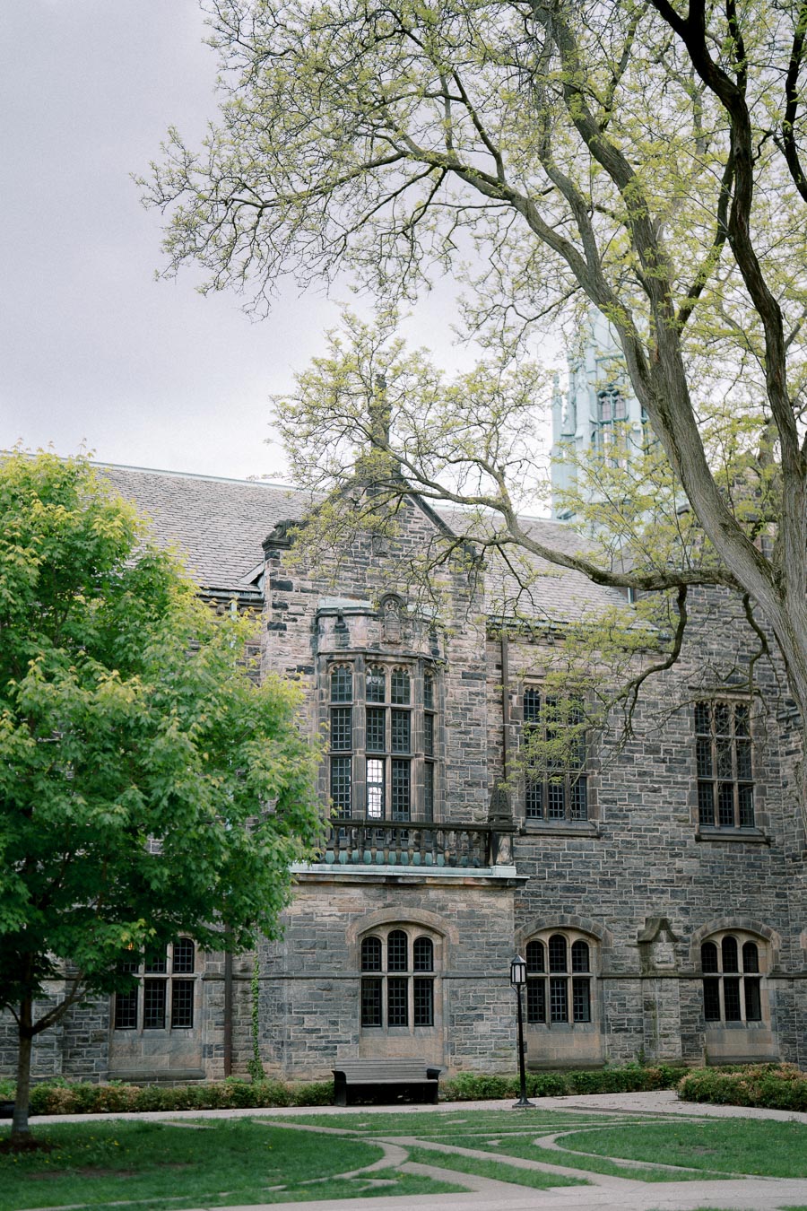 Historic stone building with large windows and trees in the foreground, set in a serene, green courtyard under an overcast sky.