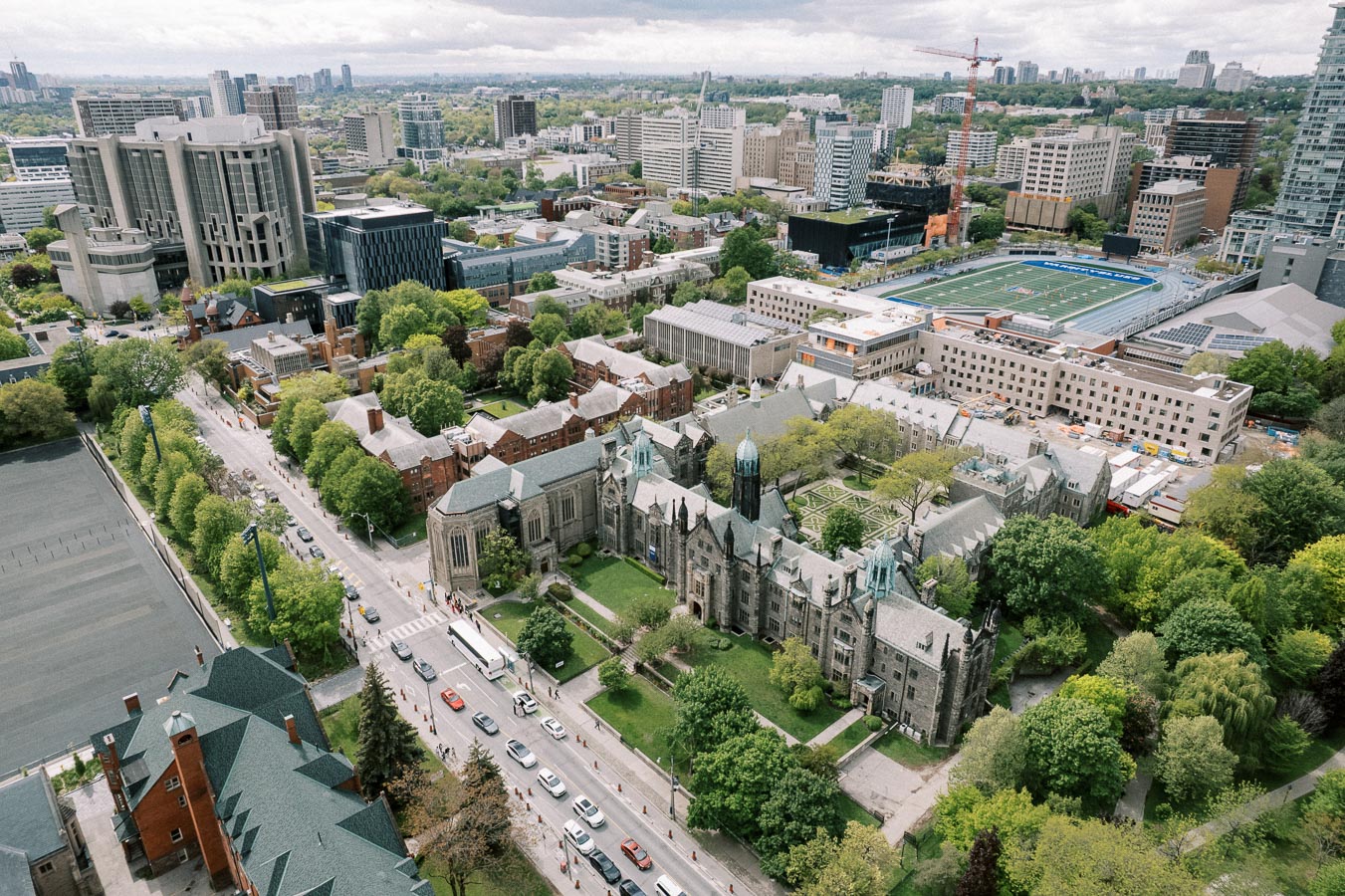 Aerial view of a lush urban campus featuring historic Gothic-style buildings surrounded by greenery and modern architectural structures, with a sports field and cityscape in the background.
