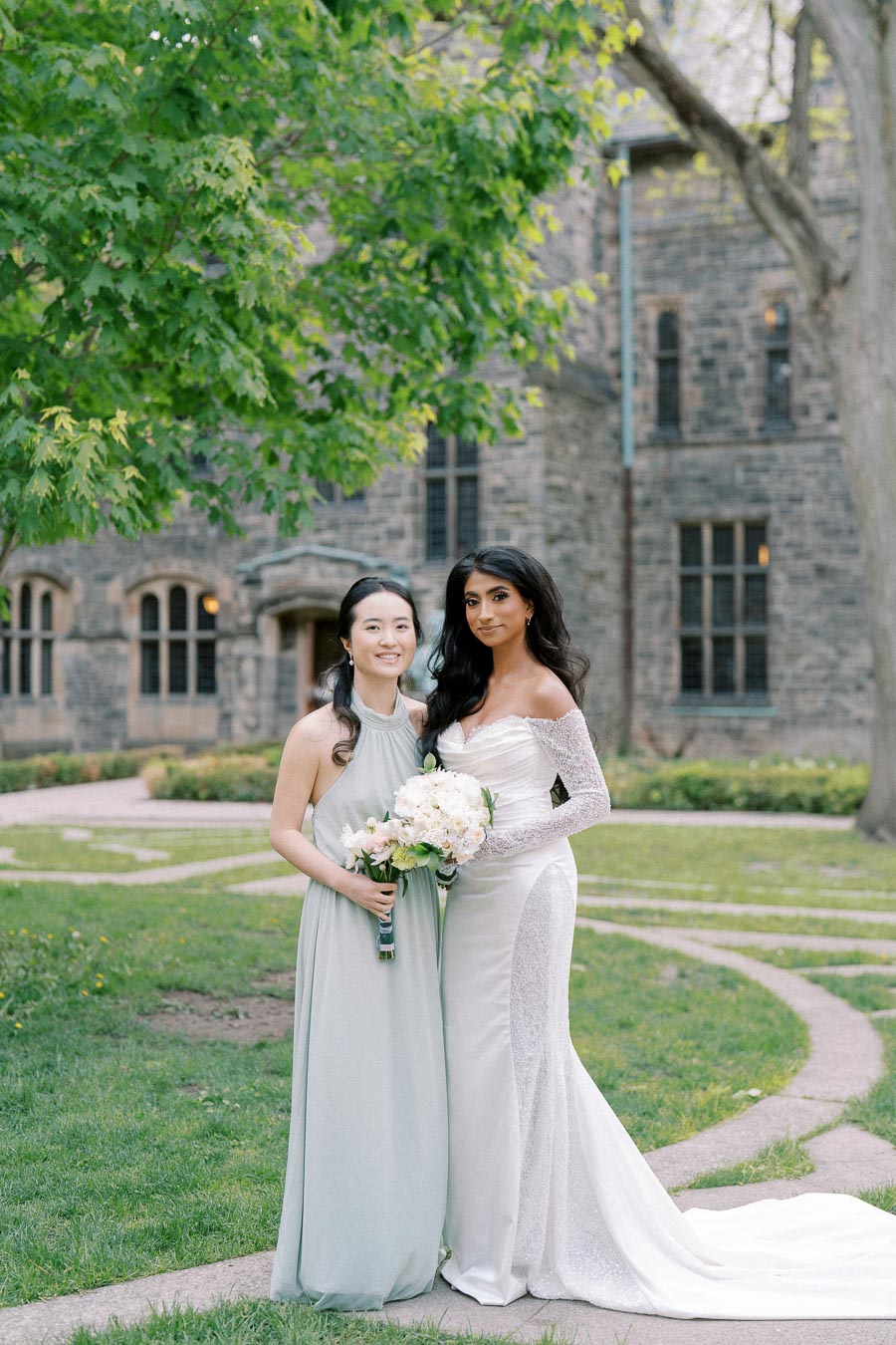 A bride in an elegant white gown poses with a bridesmaid wearing a light blue dress, both holding bouquets, outside a historic stone building with lush green surroundings.