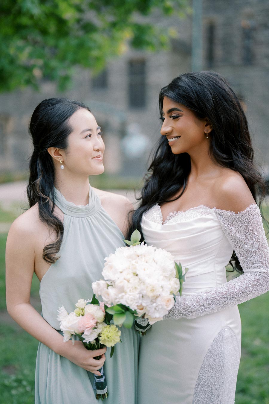 Two women at an outdoor wedding ceremony, a bride in a white gown holding a bouquet of white flowers, and a bridesmaid in a light blue dress with a bouquet of mixed flowers, smiling at each other.