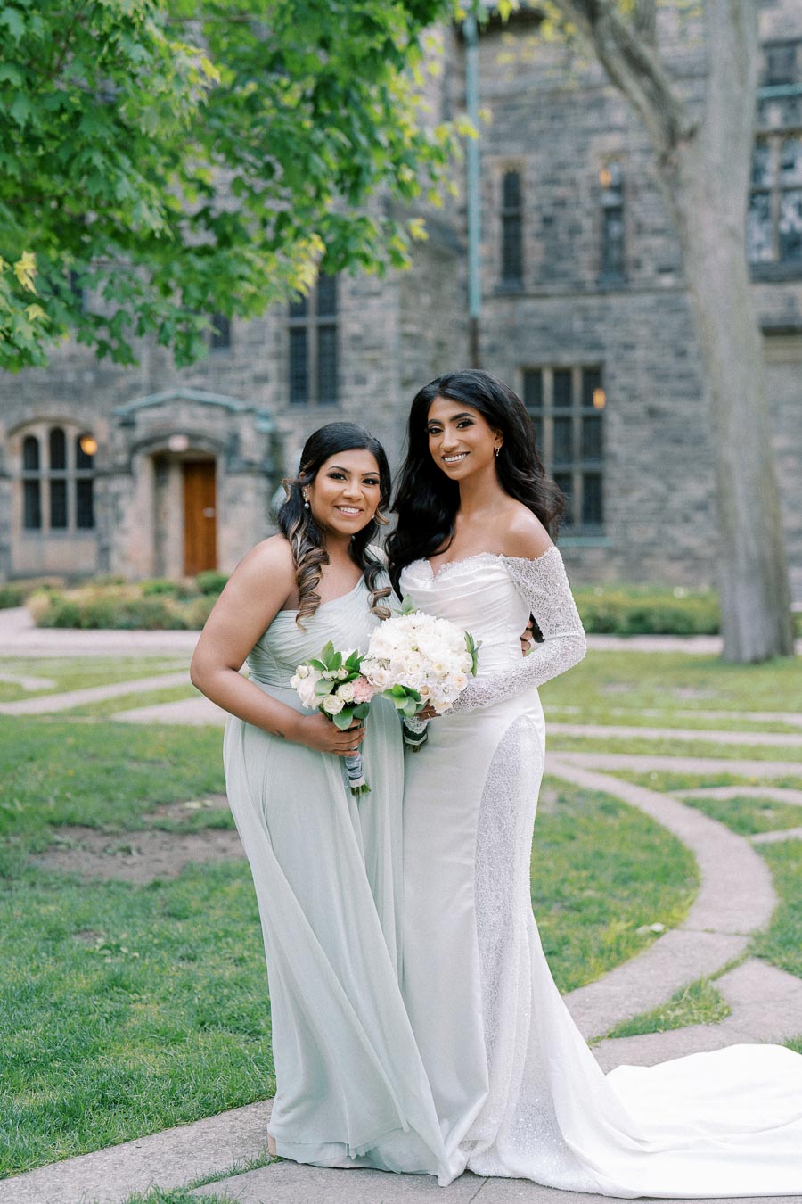 Two women in elegant dresses pose together outdoors for a wedding photo, standing on a garden path with a historic building in the background. Both women are holding bouquets, one wearing a white gown and the other a light green dress.