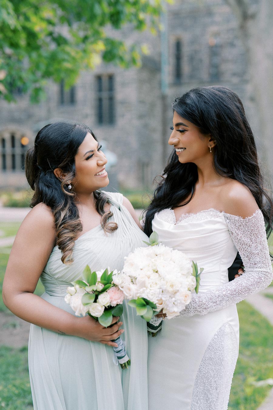 Bride and bridesmaid smiling at each other while holding bouquets, standing outdoors in front of a stone building with greenery in the background.