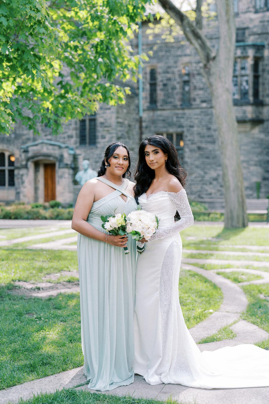 Two women in elegant dresses, one in a white bridal gown and the other in a light green bridesmaid dress, holding floral bouquets while standing together outdoors in a lush garden with a historic stone building in the background.