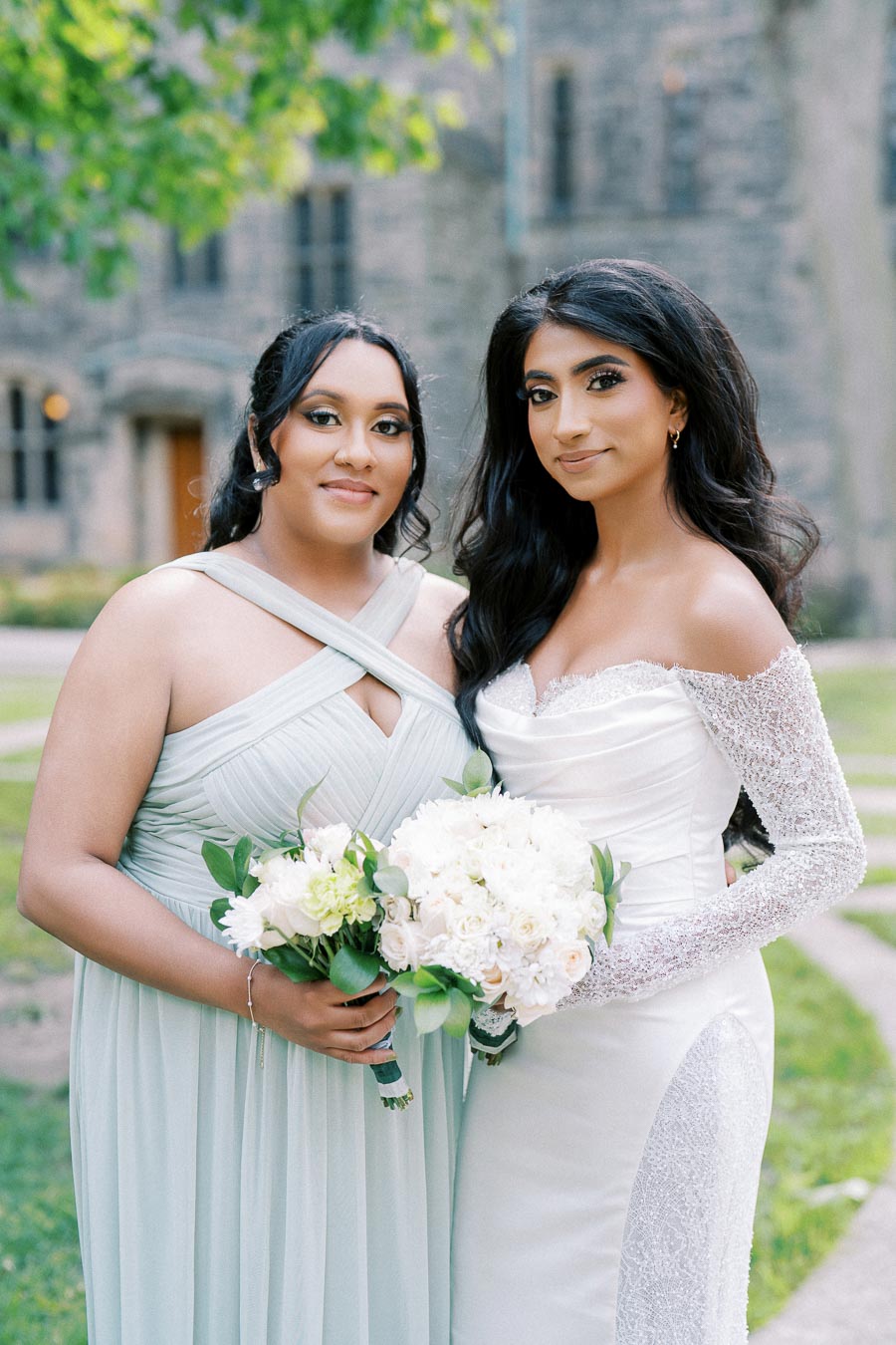 Two women posing outdoors in elegant formal dresses, holding white floral bouquets. One wears a strapless white gown, the other a light blue dress. A historic stone building and lush greenery serve as the backdrop.