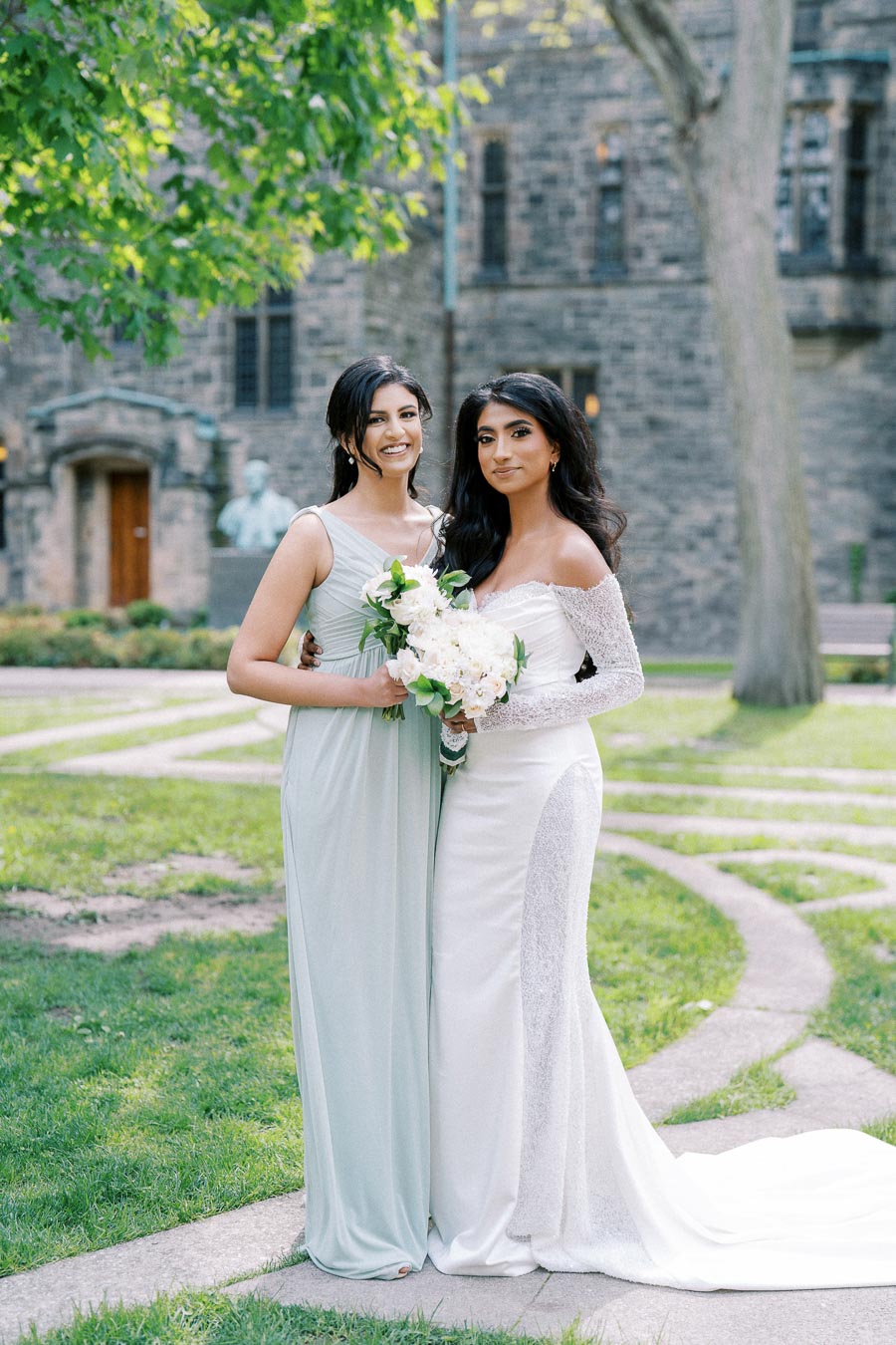 Two women posing in elegant dresses, one in white lace and one in light blue, holding a bouquet of white flowers. They stand on a manicured lawn with an old stone building and trees in the background, suggesting a wedding or formal event setting.