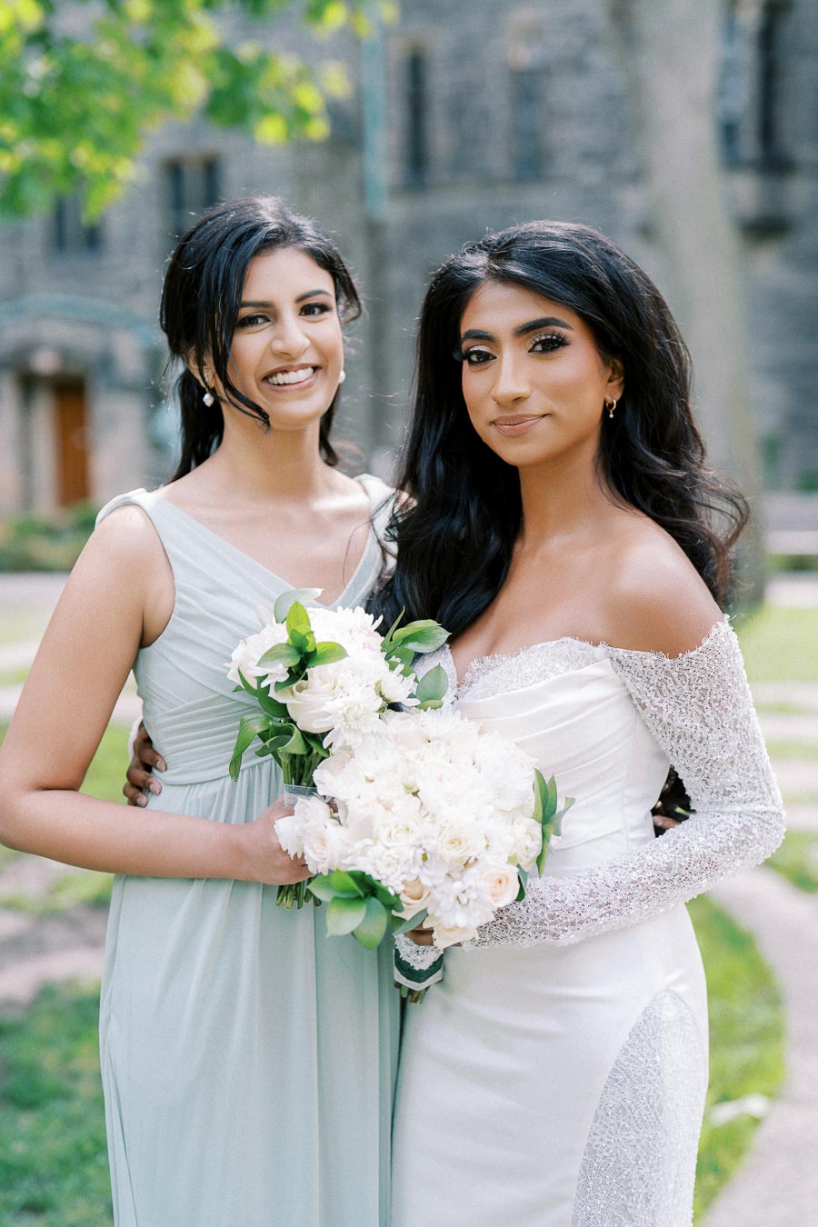 Two women dressed elegantly, one in a white bridal gown holding a bouquet of white flowers, and the other in a soft green dress, smiling outdoors with a stone building in the background.
