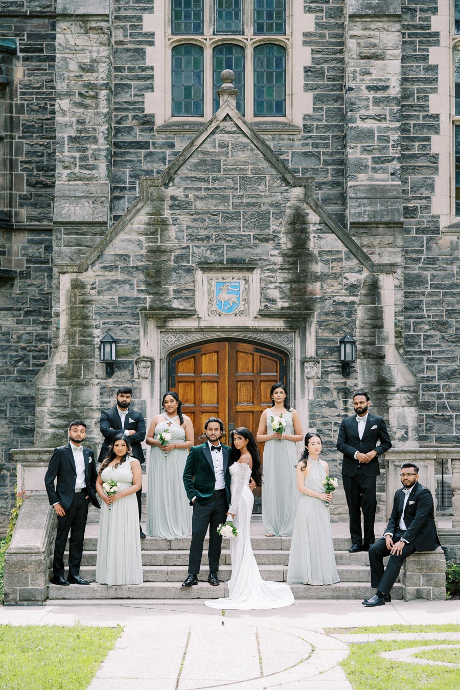 Wedding party in elegant attire posing in front of a historic stone building with arched wooden doors and emblem, featuring a bride in a white gown and groom in a dark suit, surrounded by groomsmen and bridesmaids in coordinating outfits, capturing a formal and timeless moment.
