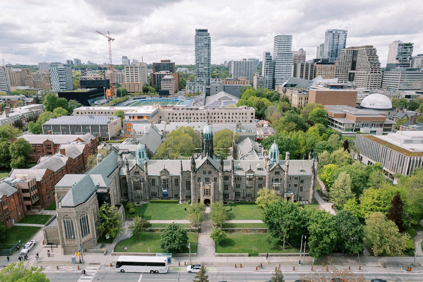 Aerial view of a university campus with historic stone buildings, surrounded by lush greenery and city skyscrapers in the background.