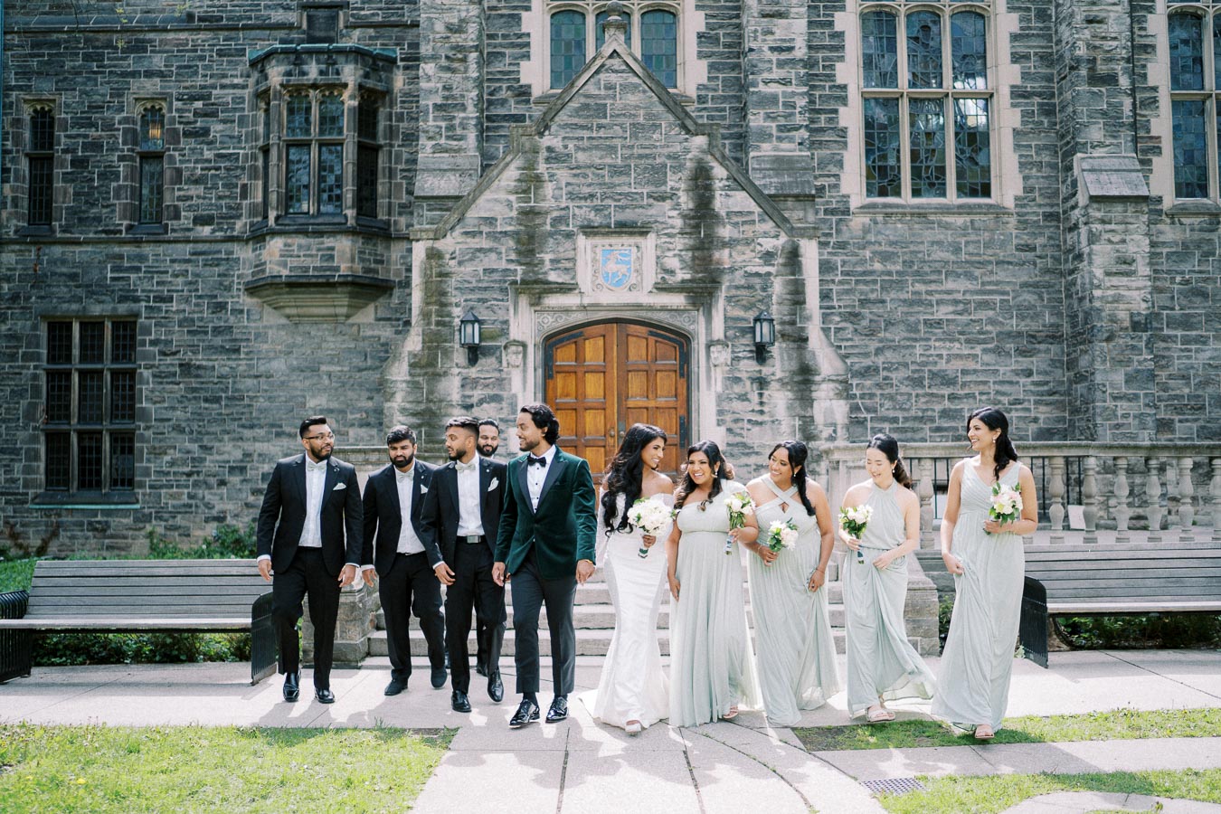 A wedding party poses in formal attire in front of a historic stone building, with groomsmen in dark suits and bridesmaids in light dresses holding bouquets.