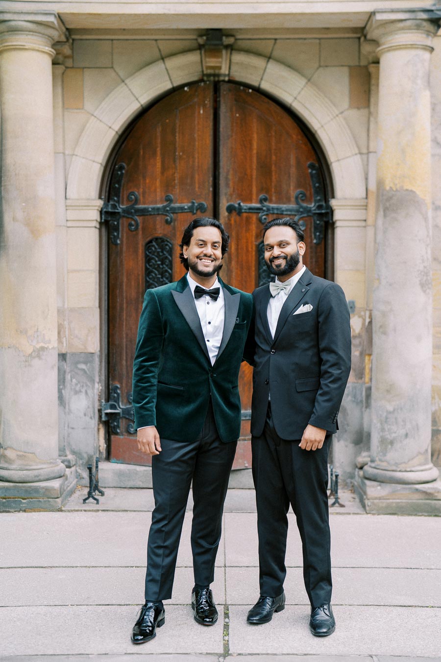 Two men in formal suits standing together in front of a large, arched wooden door with stone columns, looking directly at the camera.
