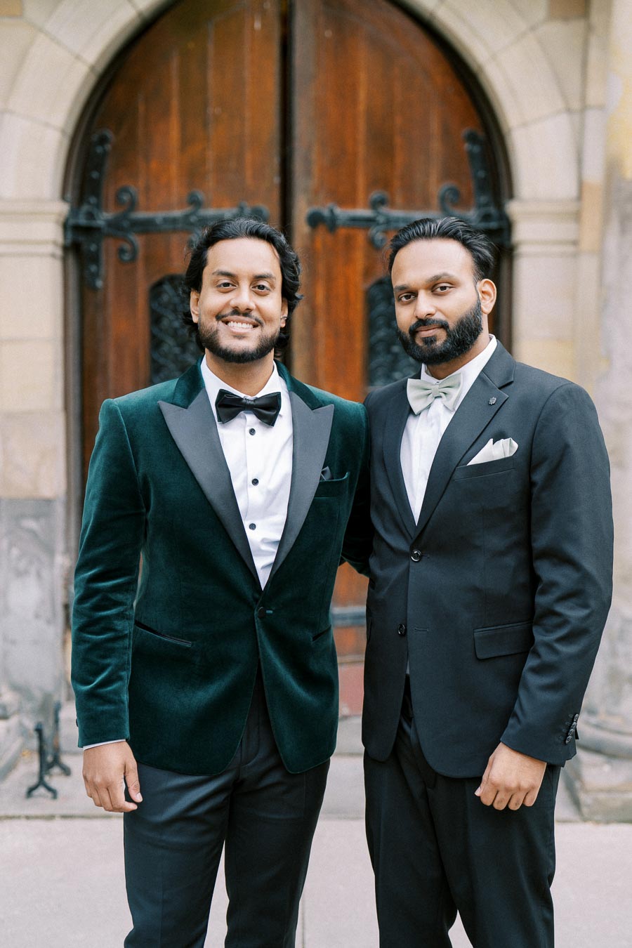 Two men in formal suits pose in front of an ornate wooden door, showcasing elegant wedding or event attire.
