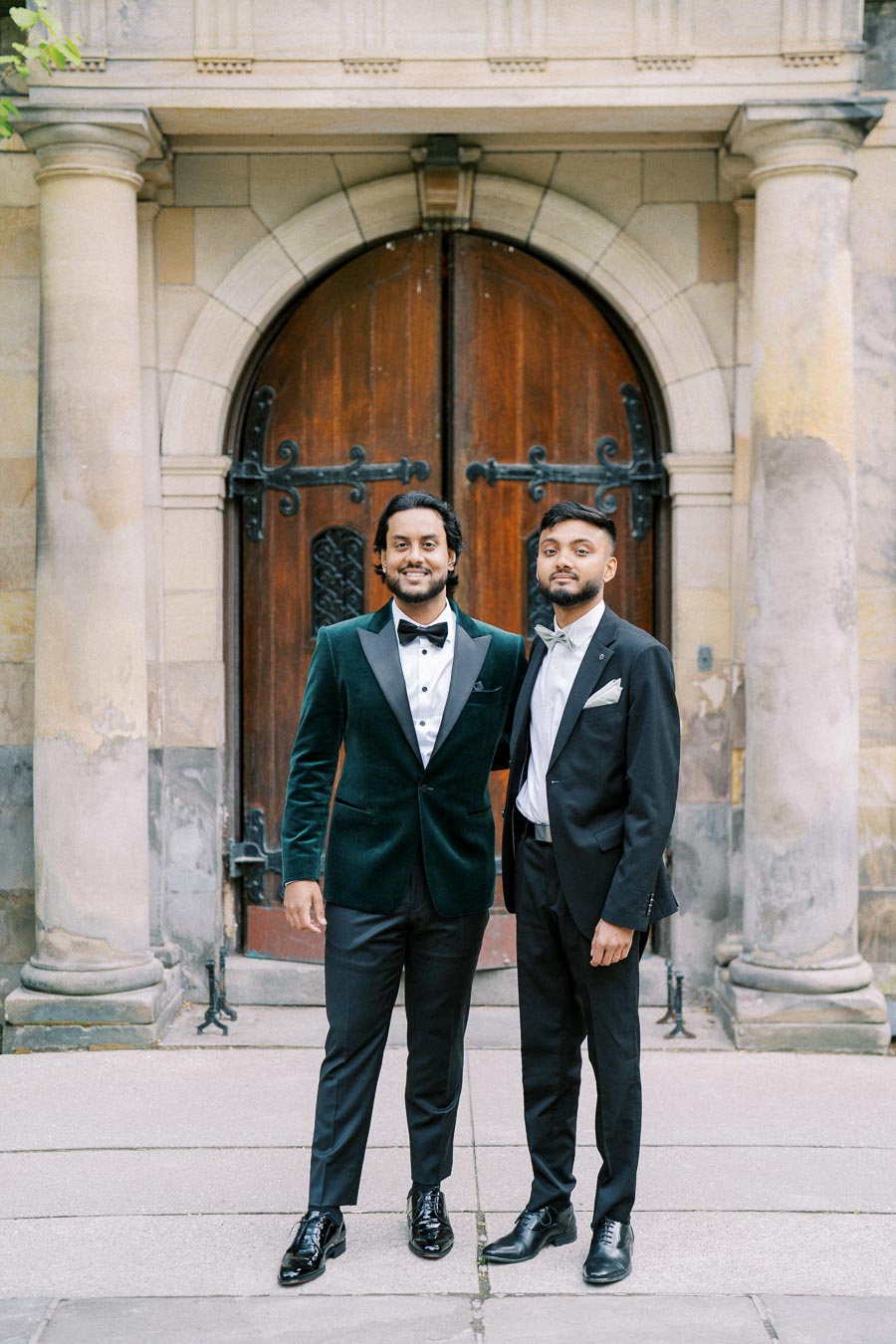 Two men in formal suits posing in front of a large wooden arched door, surrounded by stone columns, showcasing elegant wedding attire.