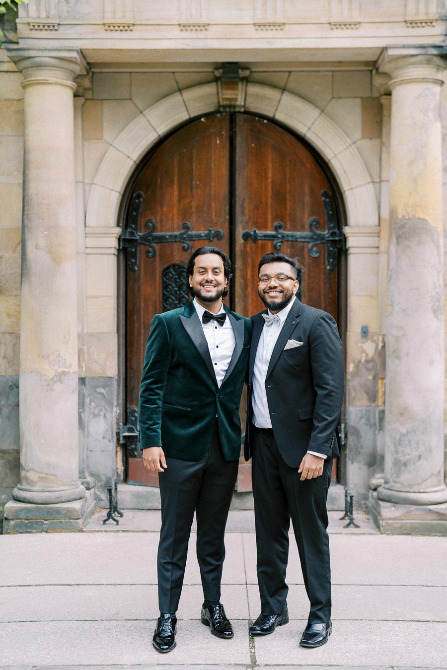 Two men in formal attire standing in front of an ornate wooden door, with stone pillars on either side, smiling at the camera outside a historic building.