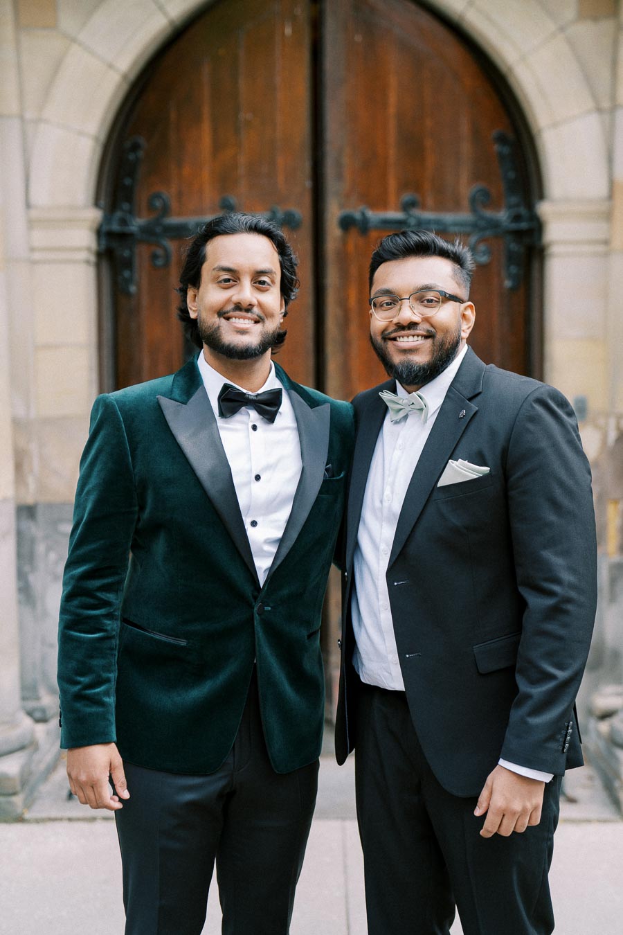 Two men dressed in formal suits with bow ties, smiling in front of a decorative wooden door at a formal event.