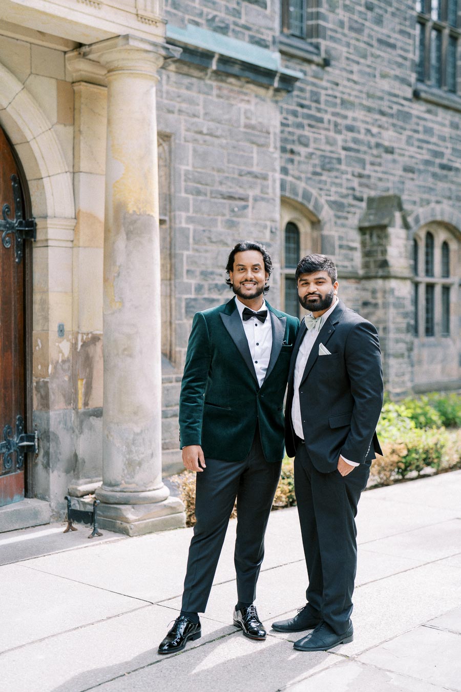 Two elegantly dressed men in suits, standing outdoors in front of a historic stone building with arched windows, exuding confidence and style.