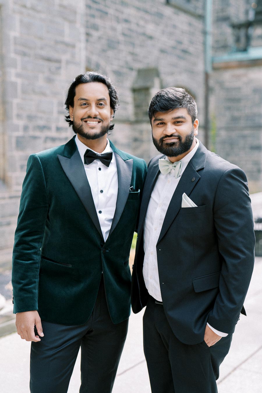 Two smiling men in formal suits, one in a velvet green jacket and the other in a black tuxedo with a bow tie, standing outside against a brick wall backdrop.