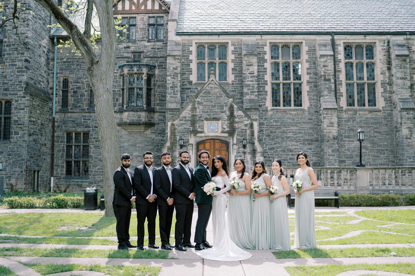 A wedding party posing in front of a historic stone building with large windows and wooden doors, featuring bridesmaids in light gray dresses and groomsmen in dark suits standing on a green lawn.