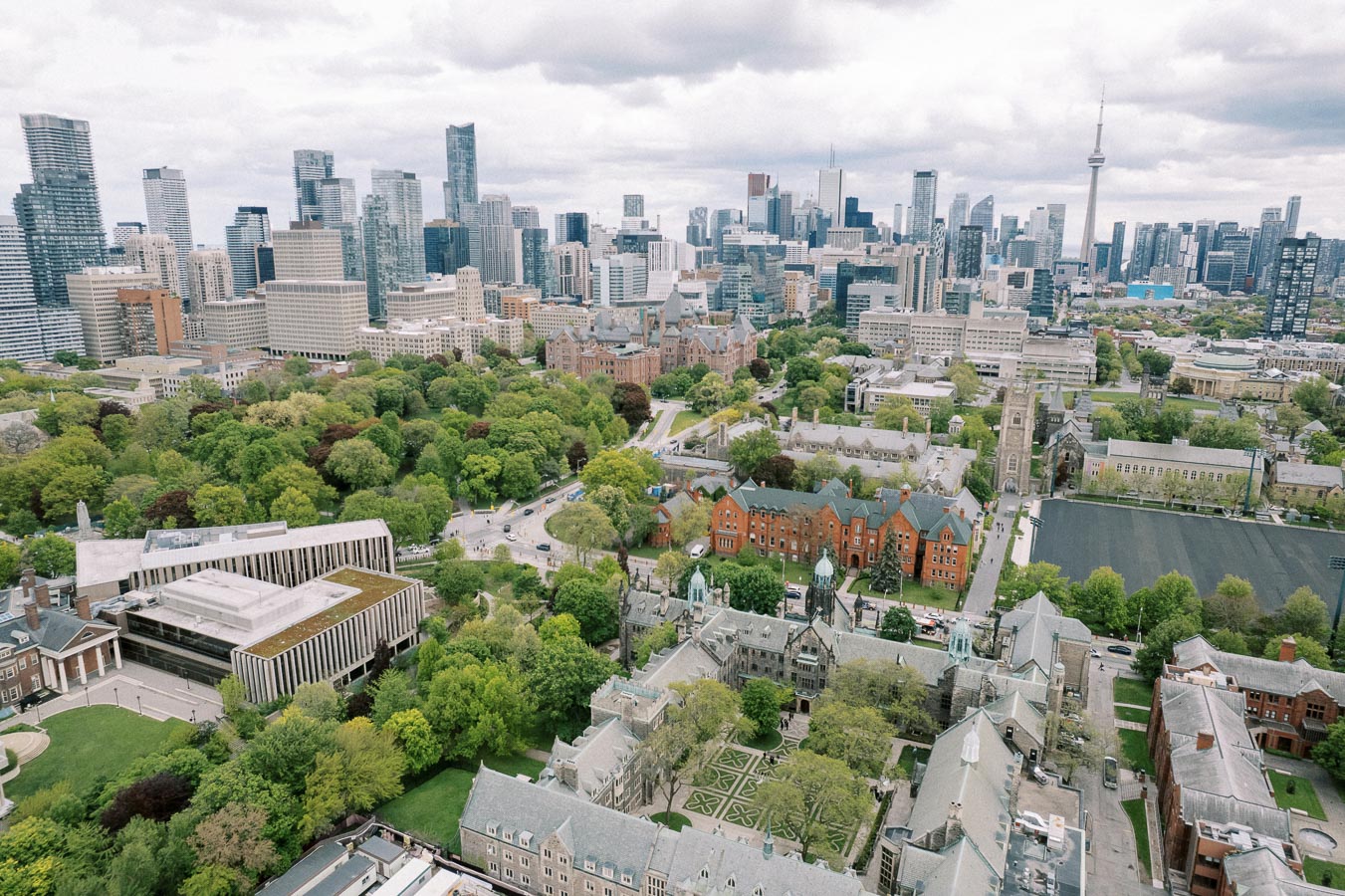 Aerial view of downtown Toronto skyline with modern skyscrapers, historic university buildings, and lush green parks under a cloudy sky.