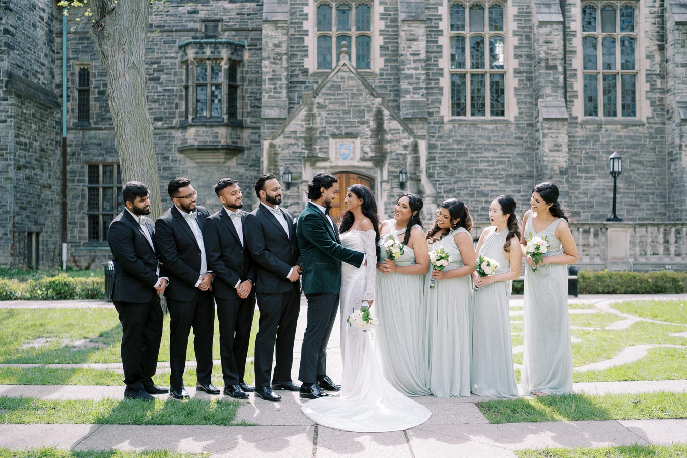 Wedding party posing in front of a historic stone building, featuring the bride in a white gown and the groom in a dark suit, surrounded by bridesmaids in pastel dresses and groomsmen in suits, all holding bouquets. The setting is outdoors on a sunny day, with greenery and elegant architecture in the background.