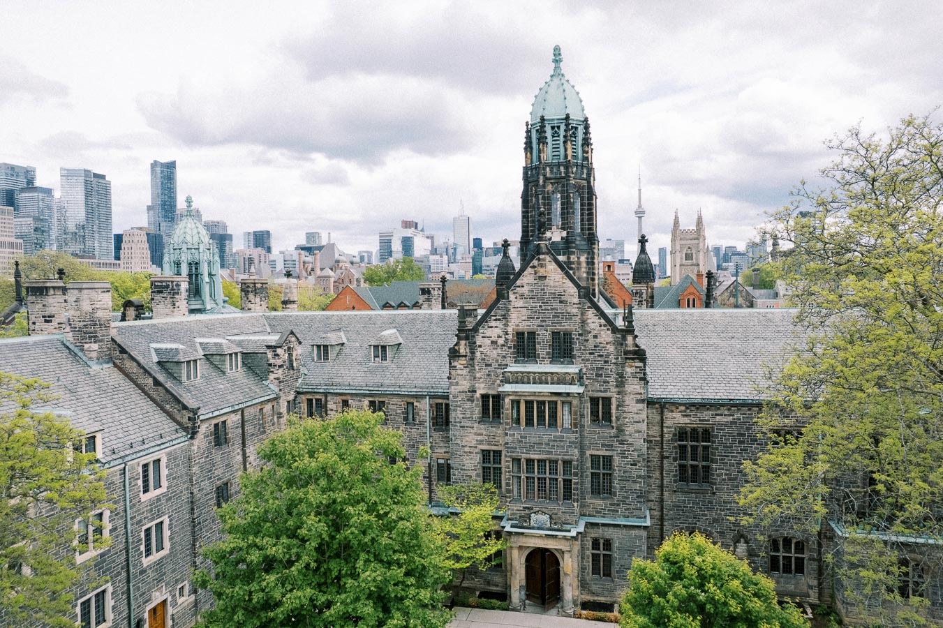 Aerial view of a historic university building, featuring a classic Gothic architectural design, with a modern city skyline in the background on a cloudy day.