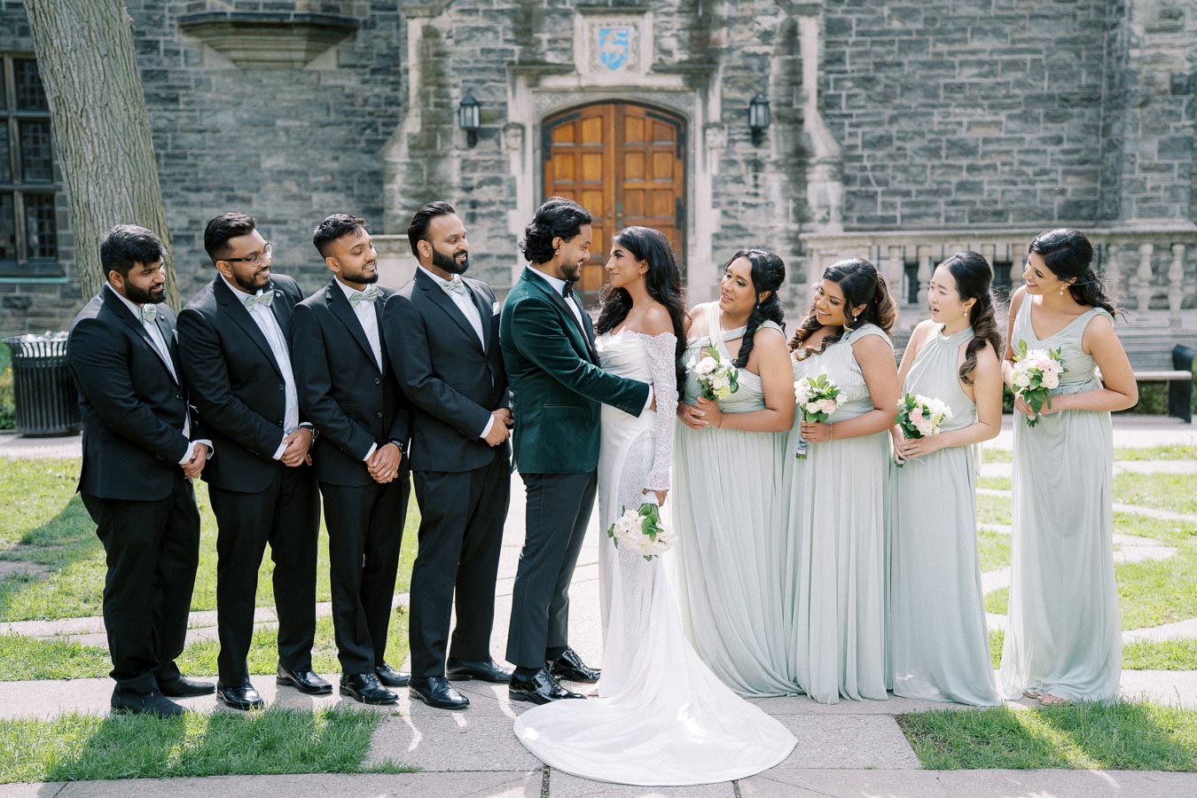 A bride and groom stand smiling with their bridal party in a formal setting. The groom wears a dark green suit, while the bride is in an elegant white dress. Their attendants include groomsmen in black suits and bridesmaids in light gray dresses holding bouquets. The backdrop is a stone building, providing a classic and timeless atmosphere.