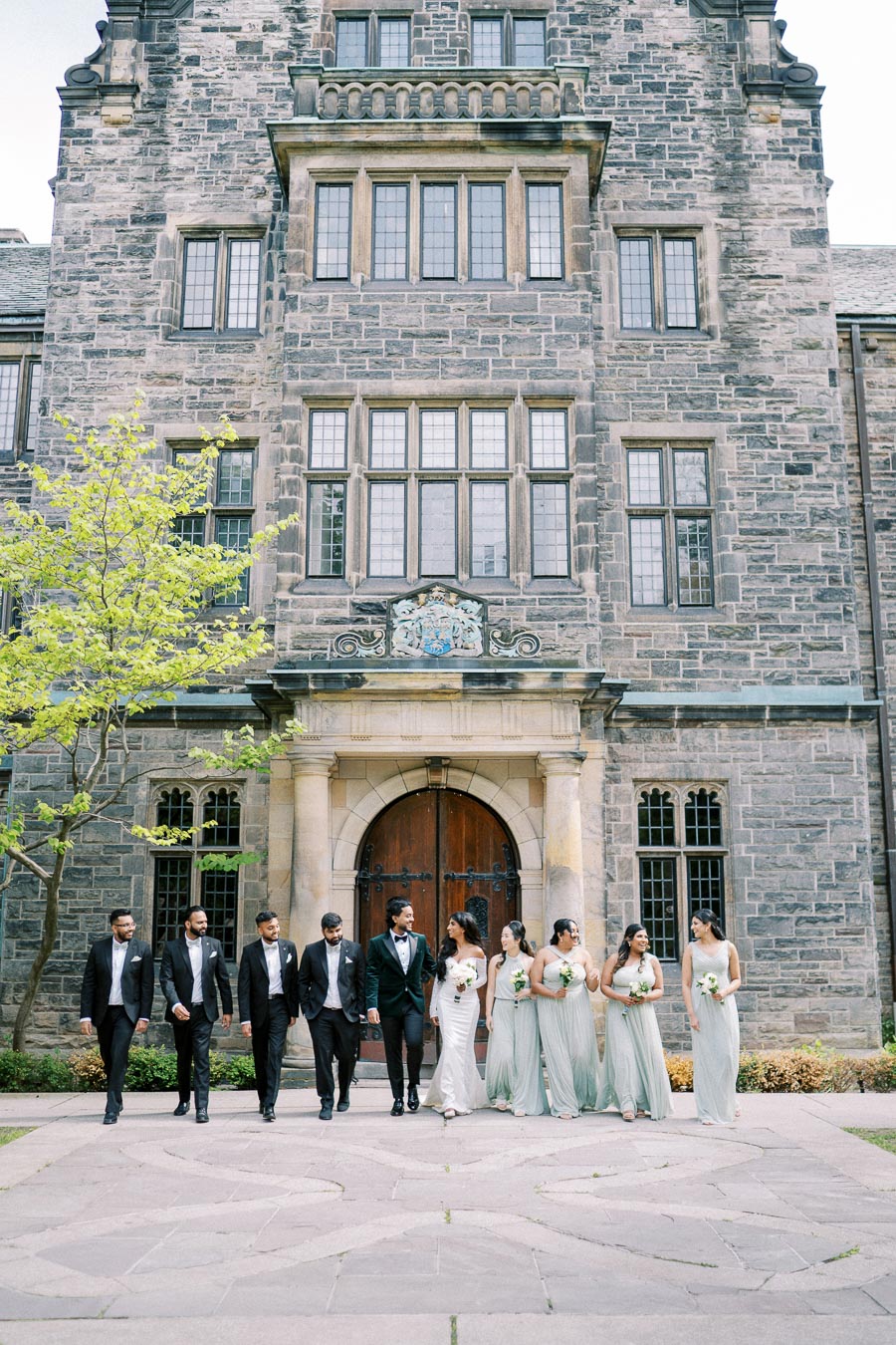 A wedding party in formal attire walking outside a historic stone building with large windows and an ornate doorway. The bridesmaids wear matching light-colored dresses, and the groomsmen are in dark suits. The scene exudes elegance and celebration.