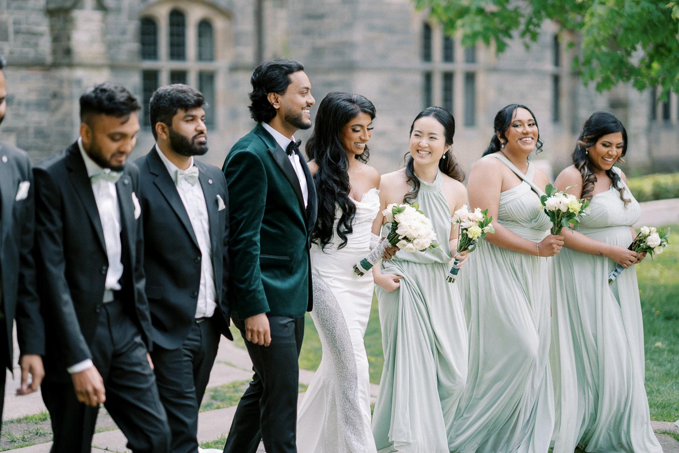 A joyful wedding party walking together outdoors, featuring bridesmaids in matching light green dresses holding bouquets, and groomsmen in black tuxedos with green velvet jackets, captured in a garden setting with a historic stone building in the background.