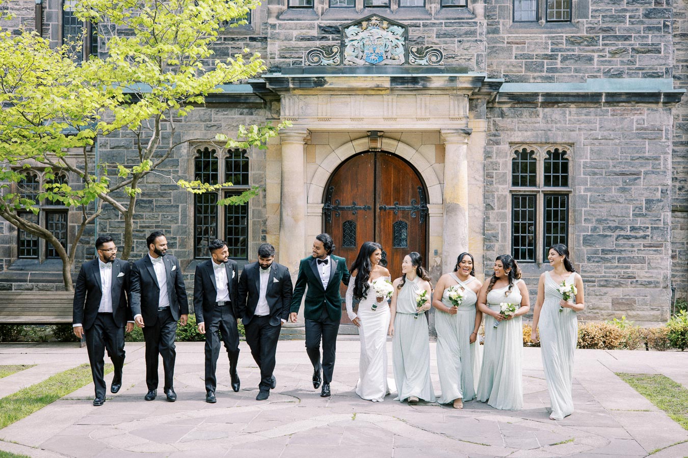 Wedding party walking outside a historic stone building, featuring groomsmen in tuxedos and bridesmaids in light gray dresses holding bouquets, under a sunny sky.
