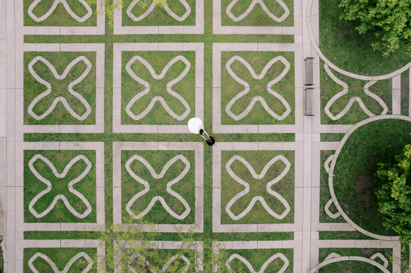 Aerial view of a geometric garden with intricate grass and pavement design. An individual with a white umbrella stands near the center, adding a focal point to the symmetrical pattern.