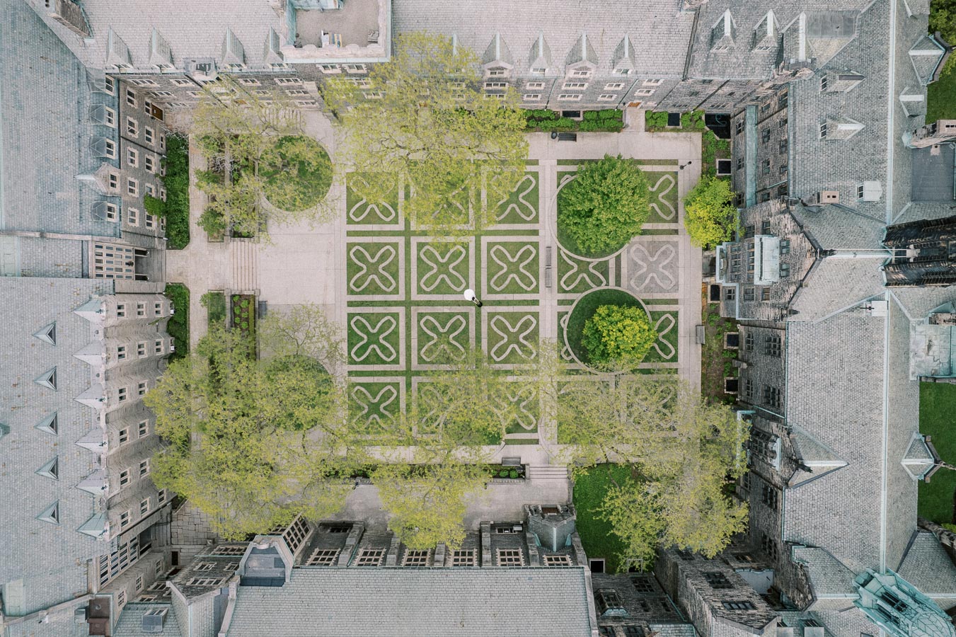 Aerial view of a historic university courtyard featuring intricate geometric gardens and surrounded by grey stone buildings with gabled roofs.