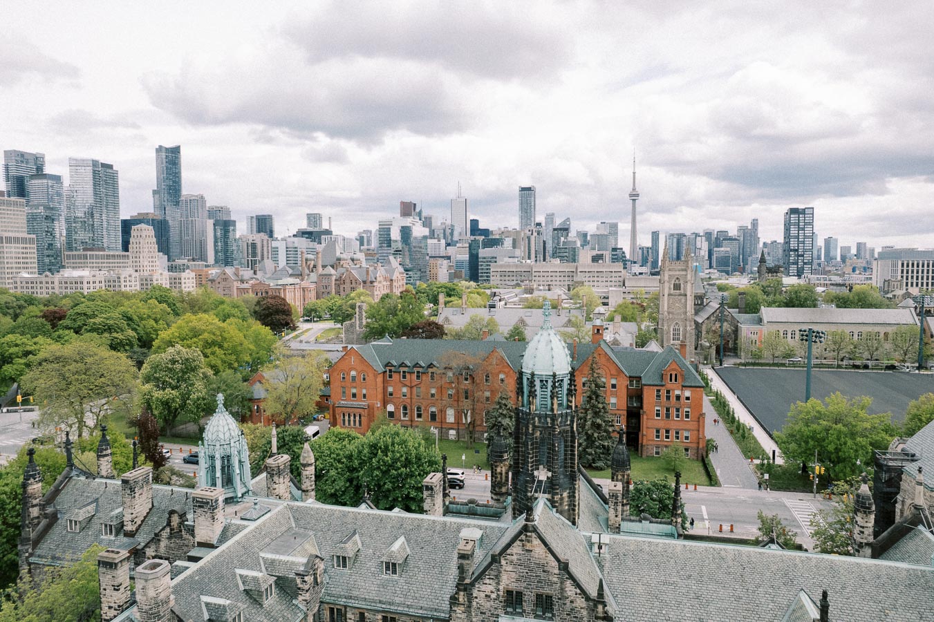Aerial view of a historic university campus with red brick buildings and lush greenery, set against a modern city skyline under a cloudy sky.