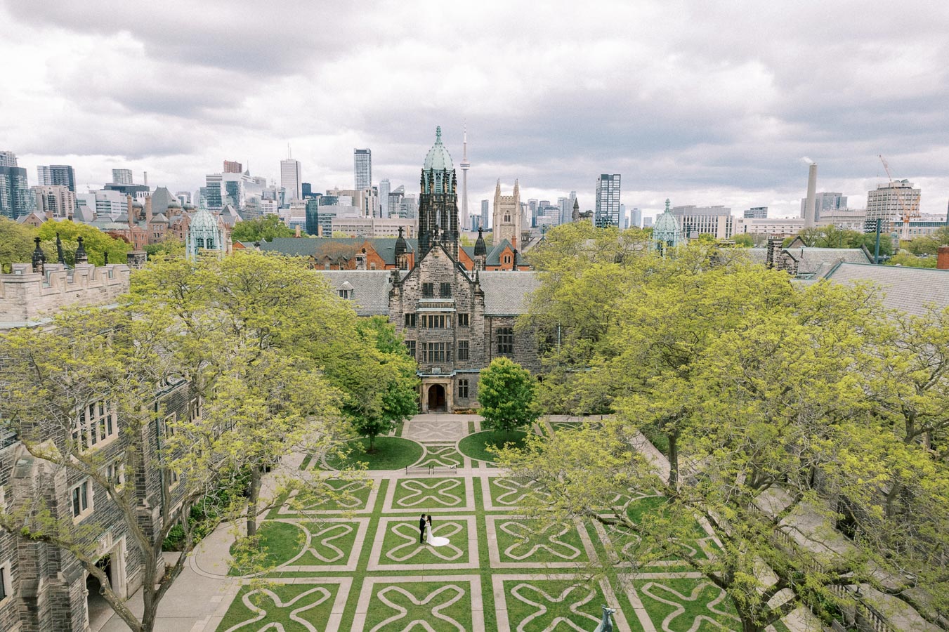 Aerial view of a historic university courtyard with intricate landscaping, surrounded by gothic architecture. The city skyline is visible in the background under a cloudy sky.