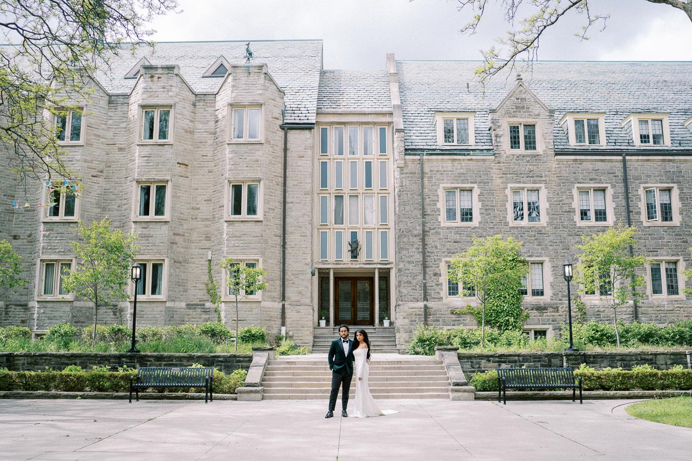 A couple in formal attire poses in front of a historic stone building with large windows and lush greenery, capturing a wedding or engagement moment on a clear day.