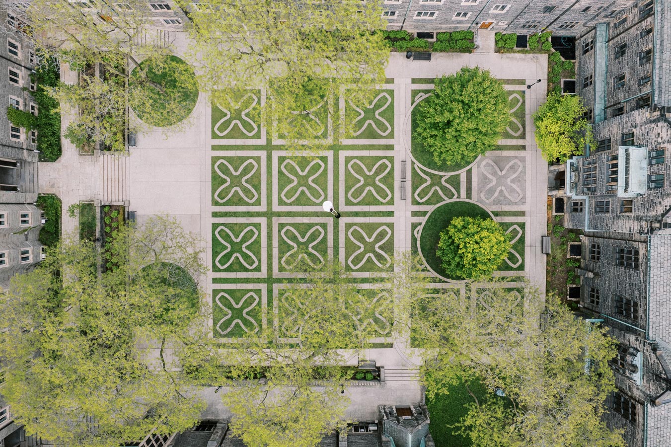 Aerial view of a beautifully designed university courtyard featuring intricate geometric patterns on the grass, surrounded by historic stone buildings and lush green trees.