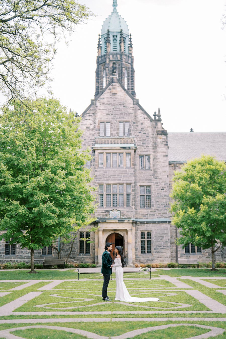 A couple in wedding attire shares a moment in front of a historic stone building, surrounded by green trees and a manicured lawn, creating a romantic and picturesque setting.