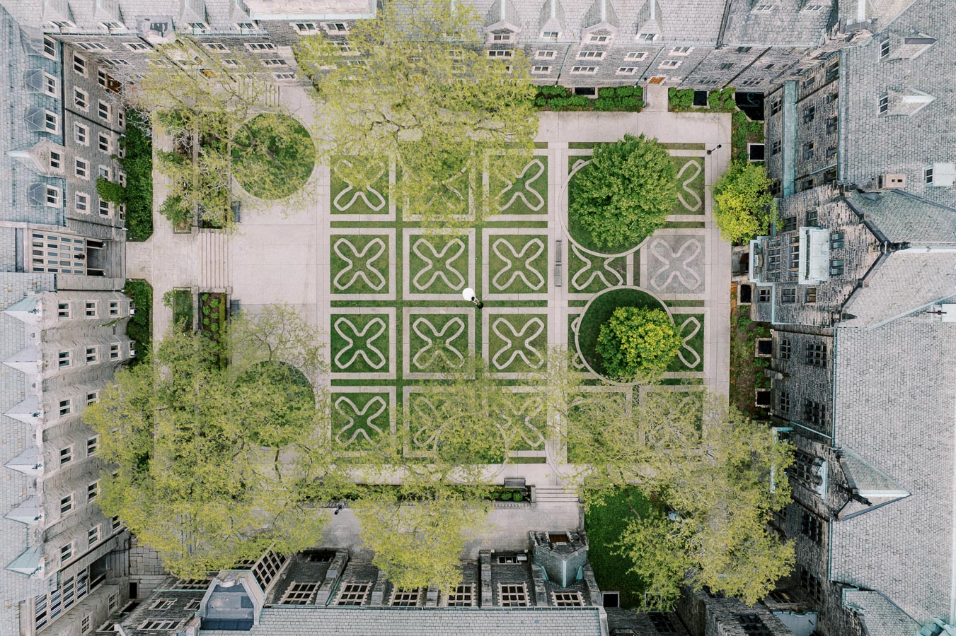 Aerial view of an intricately designed courtyard with symmetrical patterns and greenery, surrounded by historic stone buildings. The geometric layout features crisscrossing pathways and circular patches of lush trees.